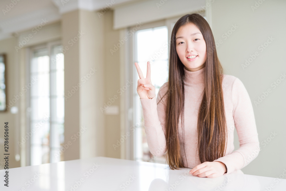 Beautiful Asian woman wearing casual sweater on white table showing and pointing up with fingers number two while smiling confident and happy. Beautiful Asian woman wearing casual sweater on white table showing and pointing up with fingers number two while smiling confident and happy.