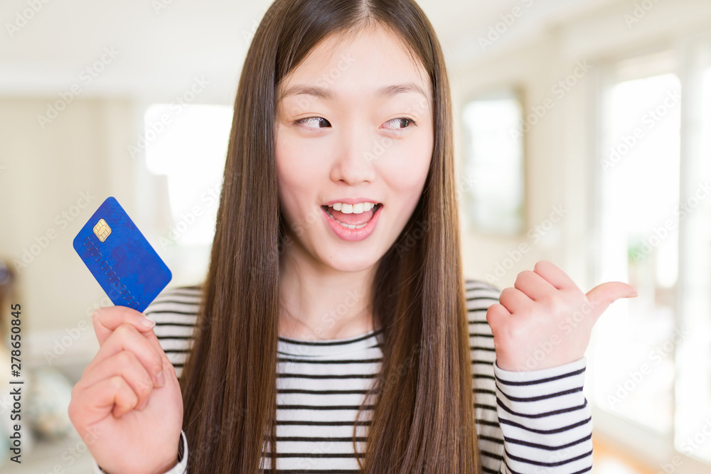 Beautiful Asian woman holding credit card pointing and showing with thumb up to the side with happy face smiling Beautiful Asian woman holding credit card pointing and showing with thumb up to the side with happy face smiling