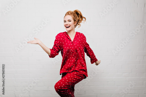 Slim positive caucasian girl in trendy red night-wear dancing on bricked background. Indoor photo of beautiful white young woman wears pajamas having fun at home.