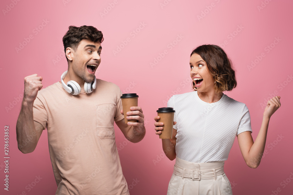 happy man and woman showing yes gestures while holding paper cups and looking at each other on pink background happy man and woman showing yes gestures while holding paper cups and looking at each other on pink background