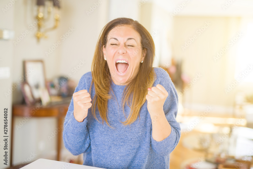 Beautiful middle age woman at home very happy and excited doing winner gesture with arms raised, smiling and screaming for success. Celebration concept. Beautiful middle age woman at home very happy and excited doing winner gesture with arms raised, smiling and screaming for success. Celebration concept.