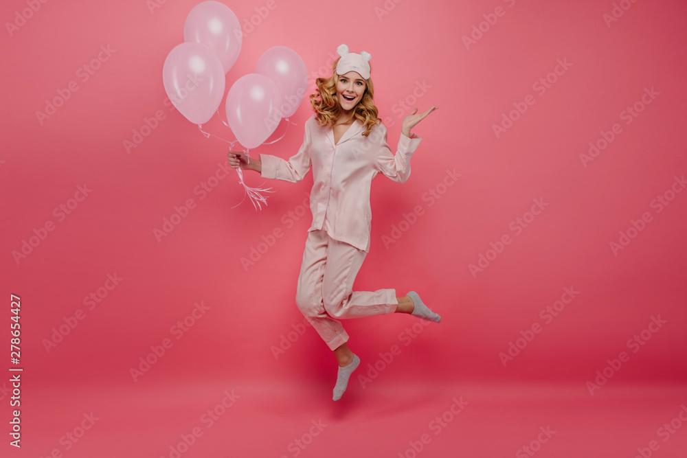 Full-length portrait of pleasant birthday girl in socks jumping on pink background. Cute young woman in pyjamas and sleepmask having fun with helium balloons.