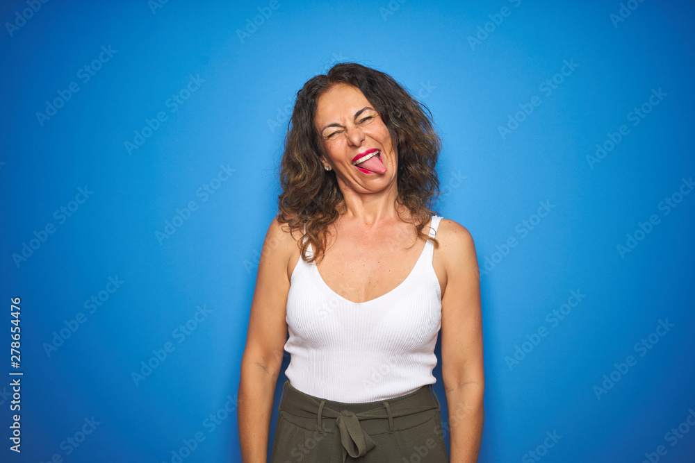 Middle age senior woman with curly hair standing over blue isolated background sticking tongue out happy with funny expression. Emotion concept. Middle age senior woman with curly hair standing over blue isolated background sticking tongue out happy with funny expression. Emotion concept.