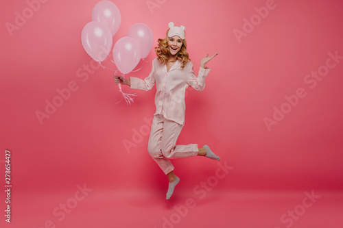 Full-length portrait of pleasant birthday girl in socks jumping on pink background. Cute young woman in pyjamas and sleepmask having fun with helium balloons.