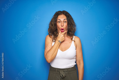 Middle age senior woman with curly hair standing over blue isolated background Looking fascinated with disbelief, surprise and amazed expression with hands on chin