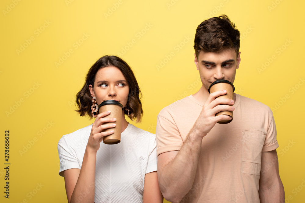 positive man and woman looking at each other while drinking coffee to go on yellow background