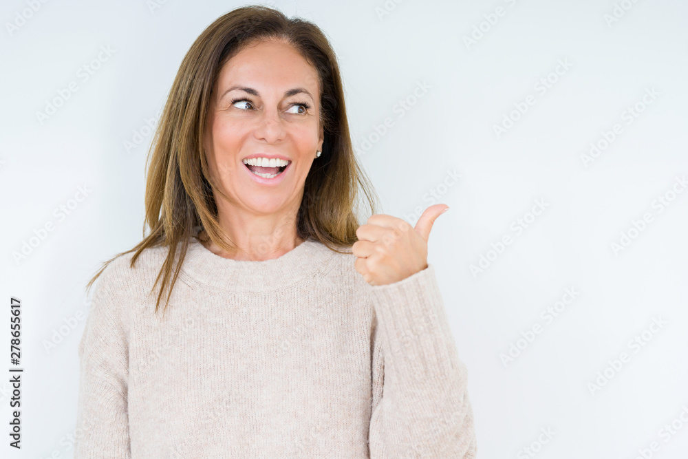 Beautiful middle age woman over isolated background smiling with happy face looking and pointing to the side with thumb up. Beautiful middle age woman over isolated background smiling with happy face looking and pointing to the side with thumb up.
