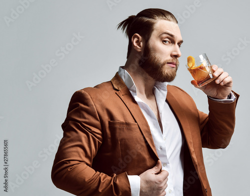Stylish modern man bearded clubber in fashion suit looks skeptically at the glass with fresh cocktail drink