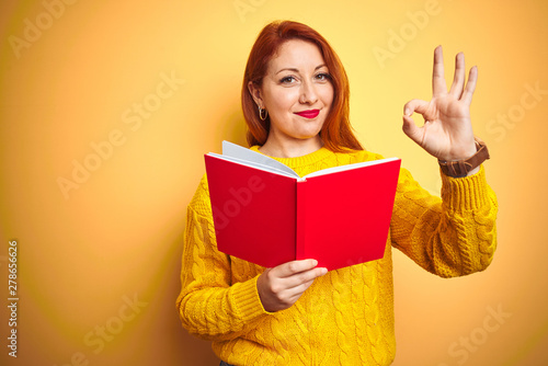 Young redhead teacher woman reading red book over yellow isolated background doing ok sign with fingers, excellent symbol