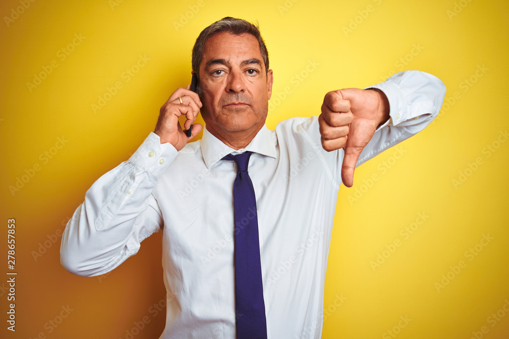 Middle age businessman talking on the smartphone over isolated yellow background with angry face, negative sign showing dislike with thumbs down, rejection concept Middle age businessman talking on the smartphone over isolated yellow background with angry face, negative sign showing dislike with thumbs down, rejection concept