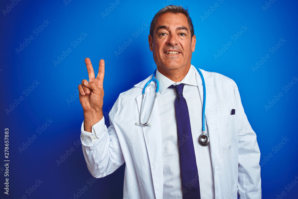 Handsome middle age doctor man wearing stethoscope over isolated blue background smiling with happy face winking at the camera doing victory sign. Number two.