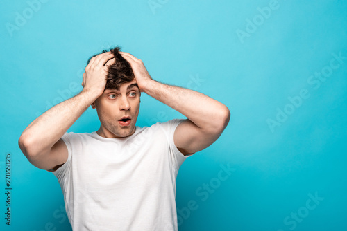 Photography shocked young man looking away while holding hands on head on blue background