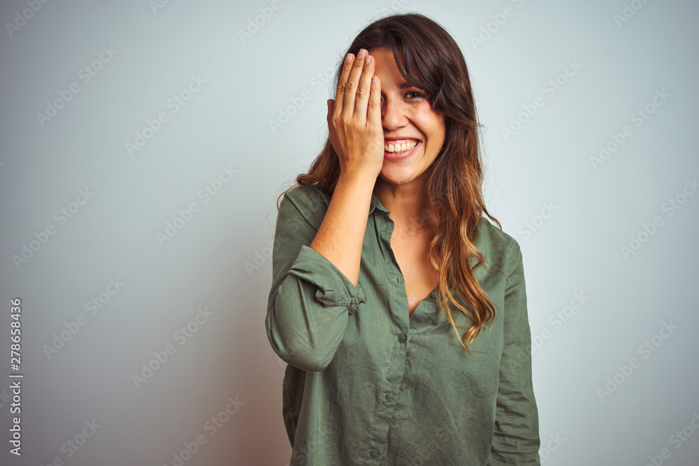 Young beautiful woman wearing green shirt standing over grey isolated background covering one eye with hand, confident smile on face and surprise emotion. Young beautiful woman wearing green shirt standing over grey isolated background covering one eye with hand, confident smile on face and surprise emotion.