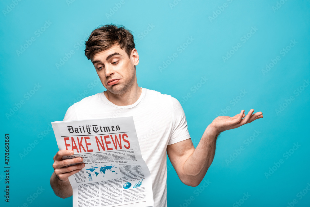 discouraged young man showing shrug gesture while reading newspaper with fake news on blue background discouraged young man showing shrug gesture while reading newspaper with fake news on blue background