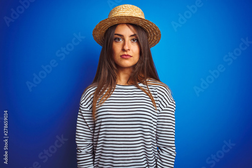 Young beautiful woman wearing navy striped t-shirt and hat over isolated blue background with serious expression on face. Simple and natural looking at the camera.