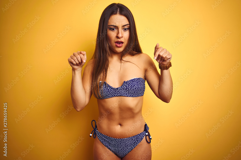 Naklejka premium Young beautiful woman on vacation wearing bikini standing over isolated yellow background Pointing down with fingers showing advertisement, surprised face and open mouth