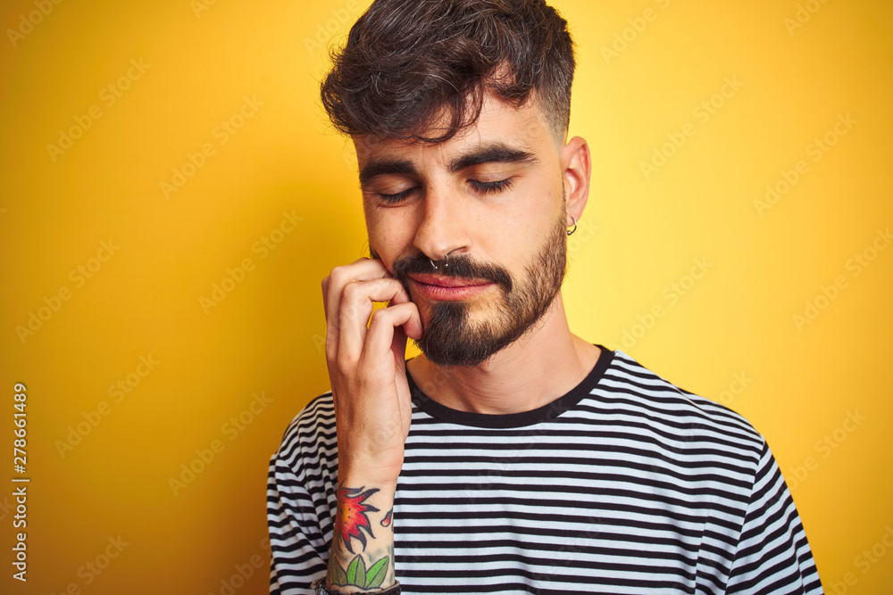 Young man with tattoo wearing striped t-shirt standing over isolated yellow background thinking looking tired and bored with depression problems with crossed arms. Young man with tattoo wearing striped t-shirt standing over isolated yellow background thinking looking tired and bored with depression problems with crossed arms.