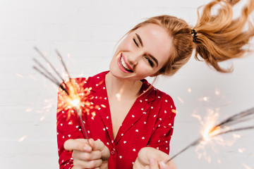 Close-up portrait of carefree girl isolated on white background with bengal lights. Studio shot of emotional lady in red night-suit posing with sparklers before new year.
