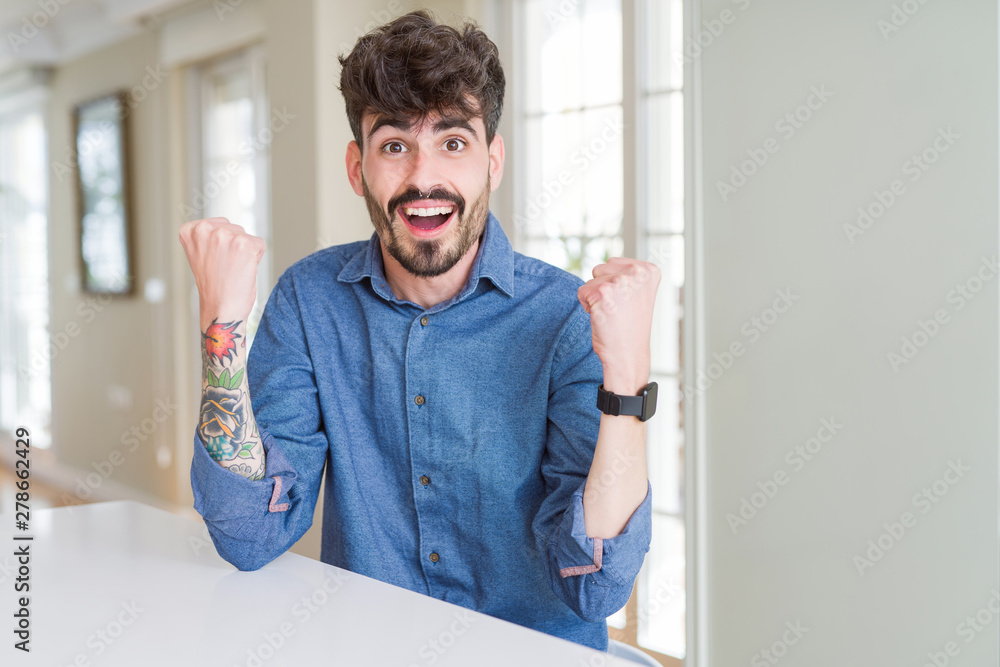 Young man wearing casual shirt sitting on white table celebrating surprised and amazed for success with arms raised and open eyes. Winner concept.