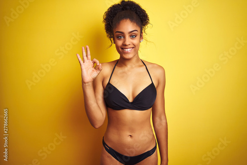 African american woman on vacation wearing bikini standing over isolated yellow background smiling positive doing ok sign with hand and fingers. Successful expression.