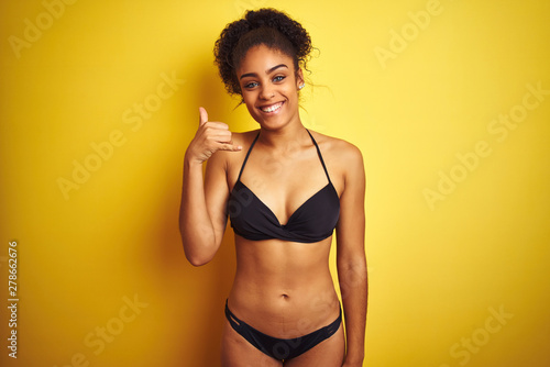 African american woman on vacation wearing bikini standing over isolated yellow background smiling doing phone gesture with hand and fingers like talking on the telephone. Communicating concepts.