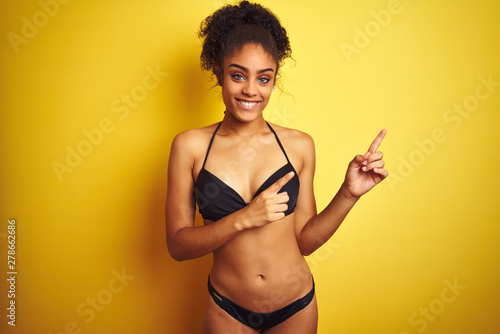 African american woman on vacation wearing bikini standing over isolated yellow background smiling and looking at the camera pointing with two hands and fingers to the side.
