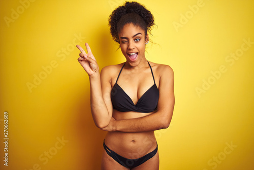 African american woman on vacation wearing bikini standing over isolated yellow background smiling with happy face winking at the camera doing victory sign with fingers. Number two. © Krakenimages.com