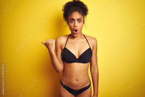African american woman on vacation wearing bikini standing over isolated yellow background Surprised pointing with hand finger to the side, open mouth amazed expression.