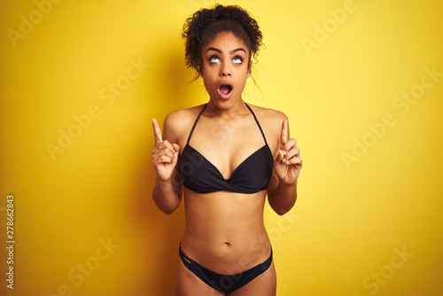 African american woman on vacation wearing bikini standing over isolated yellow background amazed and surprised looking up and pointing with fingers and raised arms.