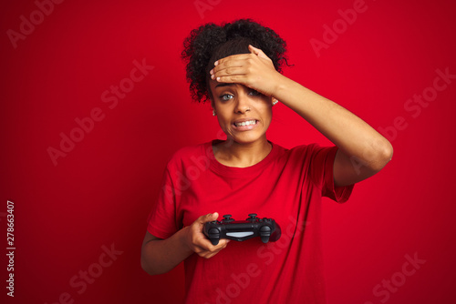 African american gamer woman playing video game using joystick over isolated red background stressed with hand on head, shocked with shame and surprise face, angry and frustrated. Fear and upset 