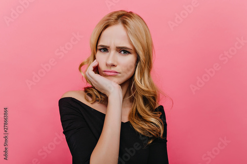 Unpleased blue-eyed lady making funny faces on bright background. Fascinating curly woman in black attire posing in studio and expressing sadness.