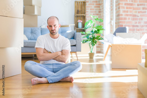 Young bald man sitting on the floor around cardboard boxes moving to a new home skeptic and nervous, disapproving expression on face with crossed arms. Negative person.