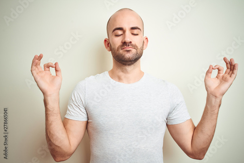 Young bald man with beard wearing casual white t-shirt over isolated background relax and smiling with eyes closed doing meditation gesture with fingers. Yoga concept.