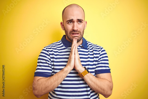 Young bald man with beard wearing casual striped blue t-shirt over yellow isolated background begging and praying with hands together with hope expression on face very emotional and worried. 