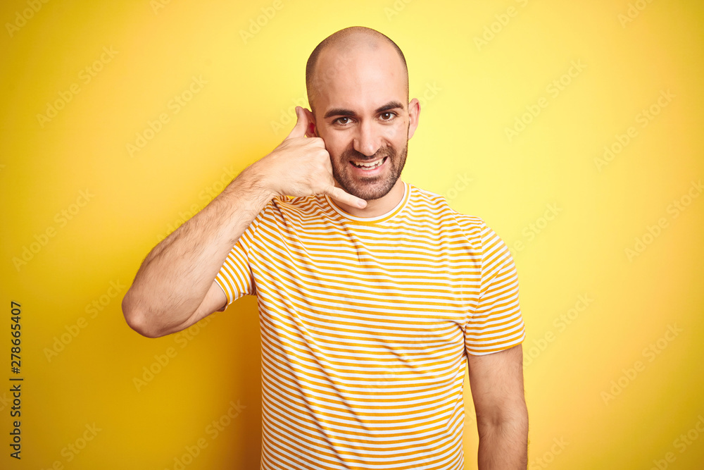 Young bald man with beard wearing casual striped t-shirt over yellow isolated background smiling doing phone gesture with hand and fingers like talking on the telephone. Communicating concepts. Young bald man with beard wearing casual striped t-shirt over yellow isolated background smiling doing phone gesture with hand and fingers like talking on the telephone. Communicating concepts.