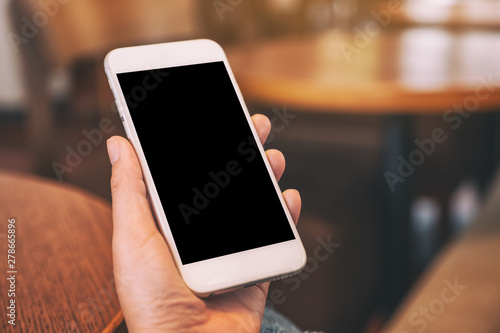 Mockup image of woman's hands holding white mobile phone with blank screen on wooden table