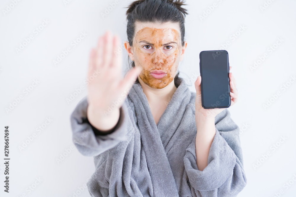 Beautiful woman wearing cosmetic facial mask as skincare treatment showing smartphone screen with open hand doing stop sign with serious and confident expression, defense gesture Beautiful woman wearing cosmetic facial mask as skincare treatment showing smartphone screen with open hand doing stop sign with serious and confident expression, defense gesture