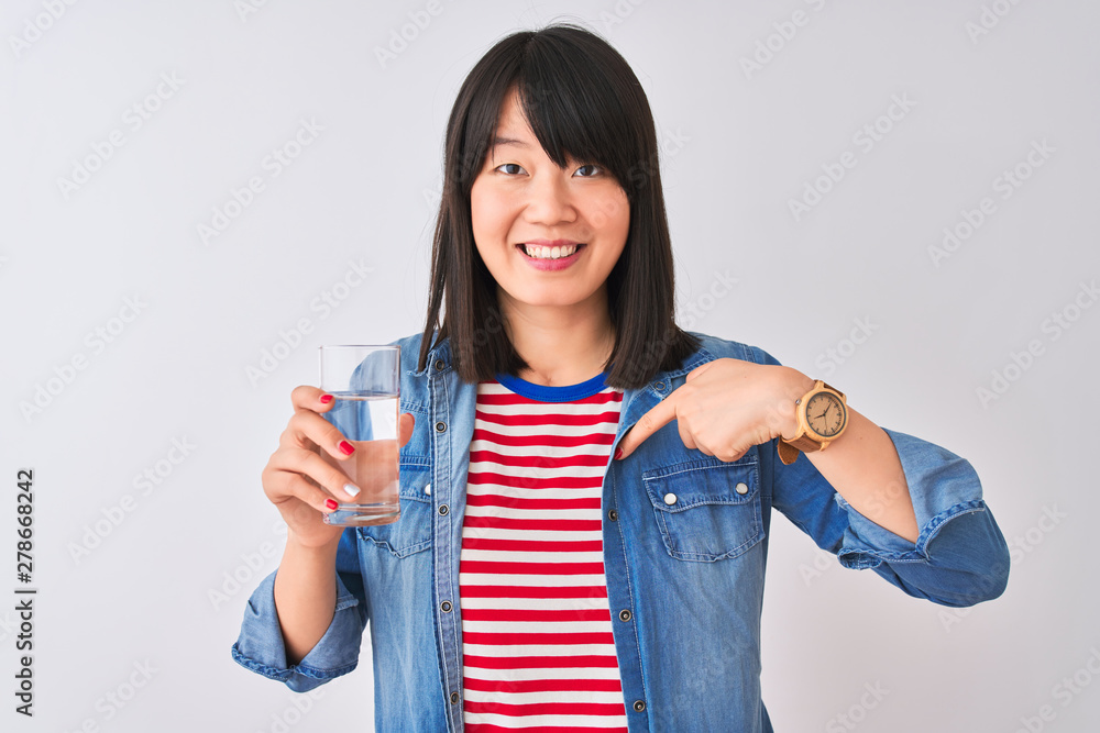 Young beautiful Chinese woman holding glass of water over isolated white background with surprise face pointing finger to himself