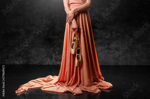 Ballerina. A young  graceful dancer standing against a black wall  dressed in a long peach dress, hands down holding pointe shoes with ribbons. Ballet studio. 3D volumetric photos.