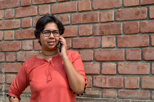Portrait of a confident looking young Indian woman with short hair and spectacles speaking on a mobile phone in outdoor setting wearing a traditional north Indian suit dress against a red brick wall
