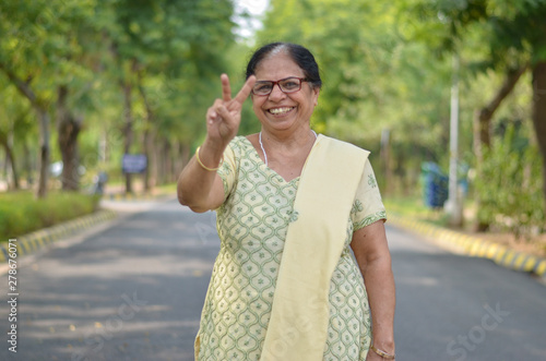 Portrait shot of a happy looking senior north Indian woman wearing traditional chikan kari Indian salwar kameez showing a Victory (V) sign in a garden against a bokeh of canopy of trees. photo