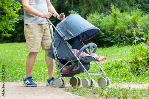 father walks with the child in park. Young man with blue stroller, front view only legs. Caring dad with summer trousers and sneakers walks with daughter, baby sleeps in stroller.