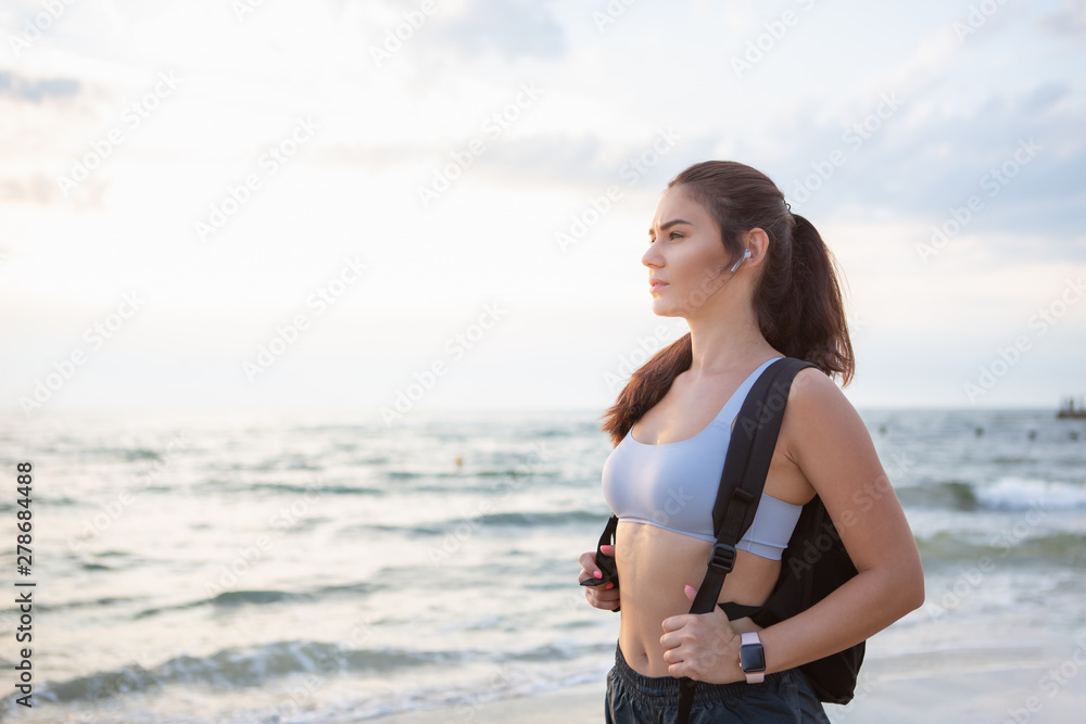 Young brunette woman with wireless earbuds, smart watches and black backpack resting after morning workout at the sea shore at sunrise. Young brunette woman with wireless earbuds, smart watches and black backpack resting after morning workout at the sea shore at sunrise.