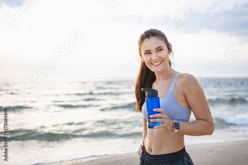 Young brunette woman with wireless earbuds and smart watches resting after morning workout at the sea shore at sunrise drinking water