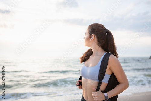 Fototapeta Naklejka Na Ścianę i Meble -  Young brunette woman with wireless earbuds, smart watches and black backpack resting after morning workout at the sea shore at sunrise.
