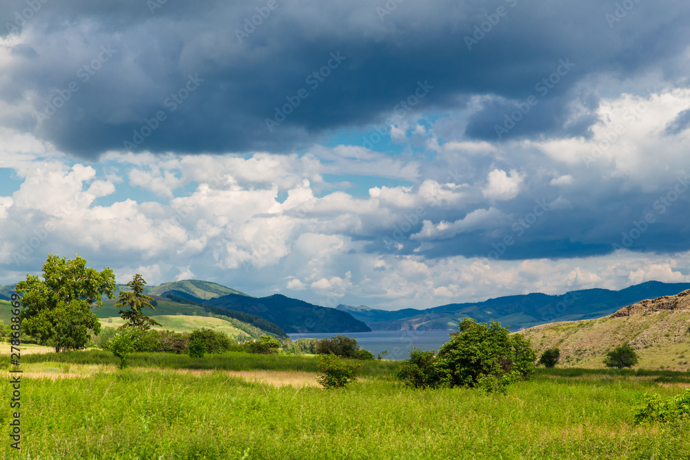 Blue sky with white clouds, trees, fields and meadows with green grass, against the mountains. Composition of nature. Rural summer landscape.