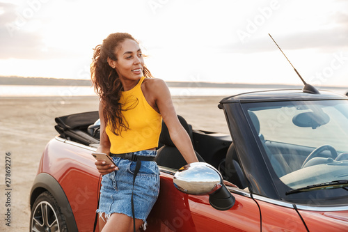 Happy smiling young pretty woman standing near car at the beach. © Drobot Dean