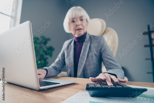 Photo of aged business lady counting sums calculator chief organization sit chair notebook table wear specs costume jacket