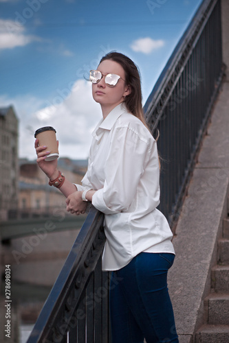 beautiful girl with take-away coffee on the background of the city and sky, craft cup, sunglasses