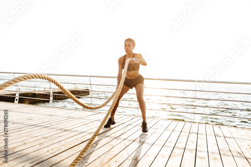 Image of athletic young woman doing workout with battle ropes by seaside in morning